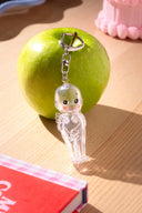 Clear Kewpie angle baby figurine keychain on a green apple with a pink cake in the background and a red book in the foreground.