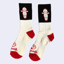 A pair of cream socks with a black color blocked part at the top with a small graphic of a Kewpie baby. The toe and the foot are red.