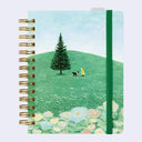 A spiral-bound notebook with a gold-tone wire binding and an illustrated cover showing a green hill, a pine tree, a small dog, and a person in a yellow raincoat. The notebook has a dark green elastic closure band and is photographed on a plain light background.