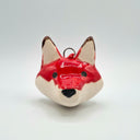 A small ceramic fox head ornament with a glossy red and cream finish, featuring black eyes and nose, set against a plain light gray background.