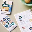 An open notebook on a counter, with journal pages filled in with Japanese kanji and stickers. Next to the notebook is a sticker sheet, with 12 different stickers within the same cohesive art style.