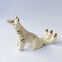 Cream colored sculpture of a sitting 3 tailed fox, with a gold poppy flower design on its back and gold tipped ears, tails and cheeks.