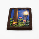 A rectangular sculpture made to look like a miniature painting within a "wooden" frame of a painted night scene, showing a glowing streetlamp, blue sky, green foliage, pink flowers, next to a wooden telephone pole, with wires hanging overhead.
