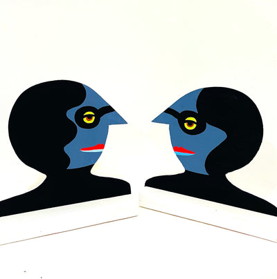 2 similar looking wooden sculptures, facing each other. Sculptures are busts with blue skin, pointed noses, black glasses and black hair.