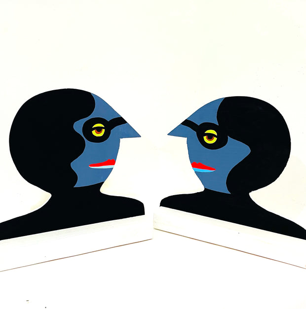 2 similar looking wooden sculptures, facing each other. Sculptures are busts with blue skin, pointed noses, black glasses and black hair.