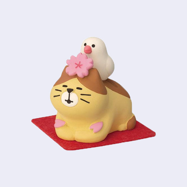 Small figure of a yellow and brown cat laying on a red mat with a cherry blossom atop its head and a white bird sitting on its back.
