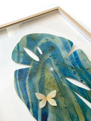 Painted cut out lily pad leaf coated in resin, blue and green with subtle marbling pattern and thin white stripes. A small gold butterfly rests atop the leaf. Displayed at an angle to show sheen of resin.
