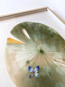Painted cut out lily pad leaf coated in resin, greyish green with subtle marbling pattern and thin black stripes. One edge of the leaf is coated in gold, as if a yellowing corner. A small blue and white butterfly rests atop the leaf. Displayed at an angle to show sheen of resin.