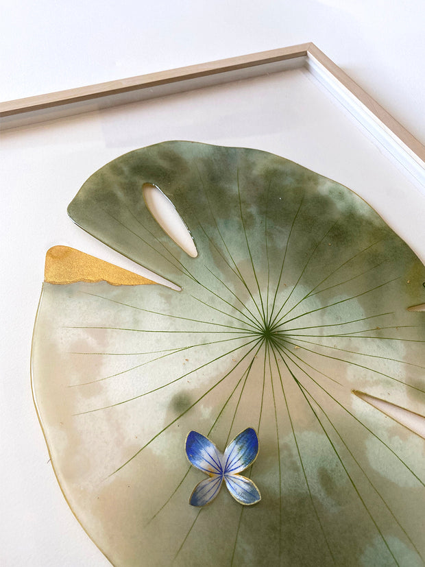 Painted cut out lily pad leaf coated in resin, greyish green with subtle marbling pattern and thin black stripes. One edge of the leaf is coated in gold, as if a yellowing corner. A small blue and white butterfly rests atop the leaf. Displayed at an angle to show sheen of resin.