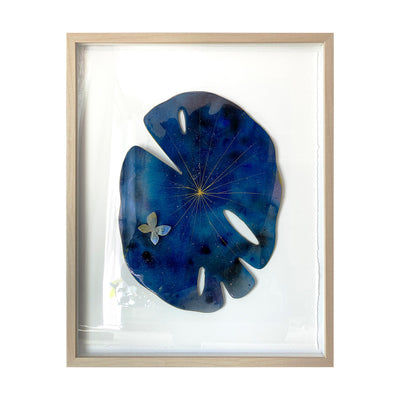 Painted cut out lily pad leaf coated in resin, dark blue with subtle galactic marbling pattern and thin gold stripes. A small blue and gold butterfly rests atop the leaf. Piece is in a thin, light grain wooden frame.