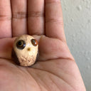 Very small sculpture of a baby owl, with brown coloring and simplistic body and face shaping. It has large bulging brown eyes and a very small yellow beak.