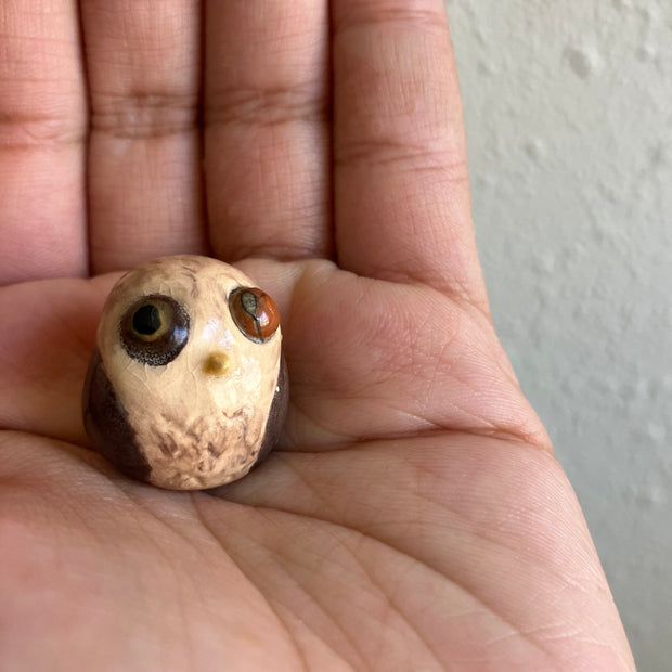 Very small sculpture of a baby owl, with brown coloring and simplistic body and face shaping. It has large bulging brown eyes and a very small yellow beak.