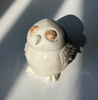 Ceramic sculpture with a crackled surface look, an off white owl with a very stout body and textured wings. It has a simple curved beak and very large eyes covered with gold glitter.