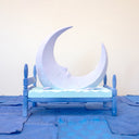 Carved wooden sculpture of a large, sharp tipped purple crescent moon with a closed eye calm expression. It rests atop of a blue bed on a dark blue wooden bed frame. Purple stars rest on the bed as well.