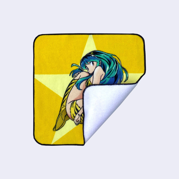 Rounded edge square towel featuring Lum, from Urusei Yatsura, slightly curled up in a ball and looking back at the viewer. The show logo is in bottom right and the background is yellow with a large yellow star. Corner folded up to show white towel backside.
