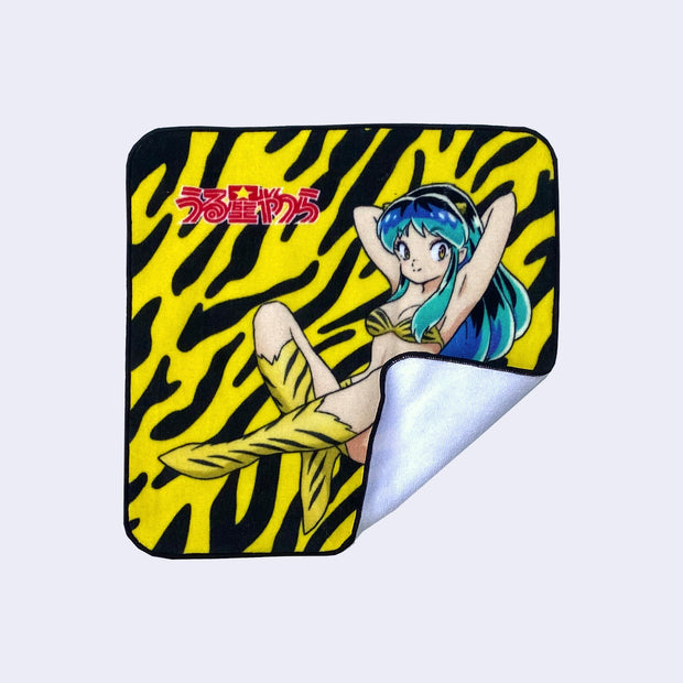 Rounded corner square towel featuring Lum from Urusei Yatsura in her classic outfit, sitting on the ground with her arms behind her head. The show's logo is in upper left and background is a yellow and black striped pattern, akin to a tiger. Corner is folded up to show white towel backside.