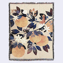 Cream color woven tapestry blanket featuring an illustration of a small woman, her back facing the viewer, sitting on a single branch of a series of large orange tree branches. Leaves are orange, dark blue and deep reddish purple and the oranges are a soft orange. 