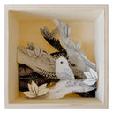 Illustrations on cut out paper, of a small greyscale hummingbird on a branch. Other branches are behind it, creating a shadow box look. Paper is outlined in gold and all contained within a thin wooden box with one open side, the viewing side.