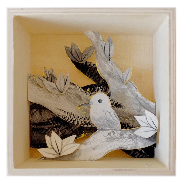 Illustrations on cut out paper, of a small greyscale hummingbird on a branch. Other branches are behind it, creating a shadow box look. Paper is outlined in gold and all contained within a thin wooden box with one open side, the viewing side.