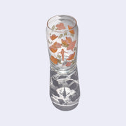 Glass cup with a flat base and slightly inward lip. A pink simply shaped woman with orange hair and no body or facial detailing stands small in the center, surrounded by same colored poppy flowers and green weed like greenery. Shown casting a stark shadow.