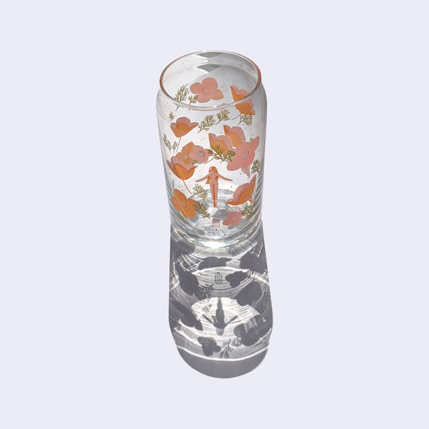 Glass cup with a flat base and slightly inward lip. A pink simply shaped woman with orange hair and no body or facial detailing stands small in the center, surrounded by same colored poppy flowers and green weed like greenery. Shown casting a stark shadow.