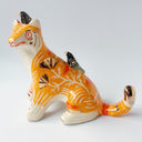 Glossy ceramic sculpture of an orange tiger with a carved pattern of poppy flowers on its body, with various stripes. Sitting on its back and head are small black magpie birds.