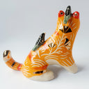 Glossy ceramic sculpture of an orange tiger with a carved pattern of poppy flowers on its body, with various stripes. Sitting on its back and head are small black magpie birds.