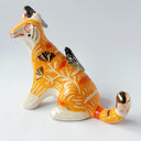 Glossy ceramic sculpture of an orange tiger with a carved pattern of poppy flowers on its body, with various stripes. Sitting on its back and head are small black magpie birds.