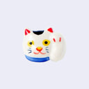 3D ceramic brooch/pin of a maneki cat's head with one paw raised upward. It has a smile, yellow eyes and a blue collar.