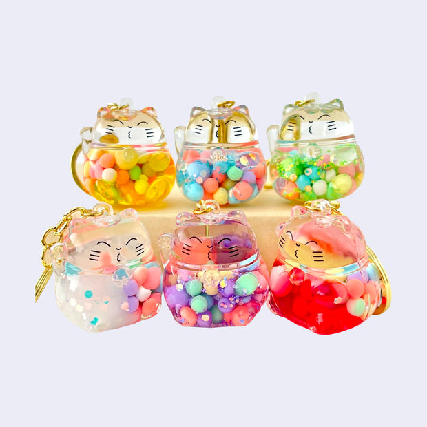 Clear plastic keychains shaped like round maneki cats, each with colorful balls, liquid and glitter within them. Each cat has a cute drawn on face.