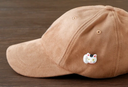 3D ceramic brooch/pin of a maneki cat's head with one paw raised upward. It has a smile, yellow eyes and a red collar and is shown on a brown cap.