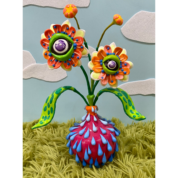 Very colorful semi psychedelic inspired sculpture of a hot pink plant bot with blue water droplets running out of it. A plant with 2 flower buds has eyes for their centers, coming out of the pot.