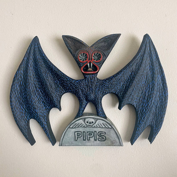 Wood sculpture of a bat, black with blue fur and a frightening face, red eye and lip outline. It stands with its wings spread atop of a half circle grave stone that reads "Pipis"