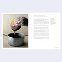 Hand with tattoo dipping a donut into a pot of chocolate sauce on a textured surface.
