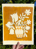 Illustration of floral design and vase on a yellow background, held against a green leafy background.