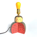 Lamp without a shade and a ceramic base of a sliced open watermelon, with a person's head on top. They have a simple displeased expression and have their lightbulb atop their head.