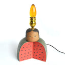 Lamp without a shade and a ceramic base of a sliced open watermelon, with a person's head on top. They have a simple stoic expression and have their lightbulb atop their head.