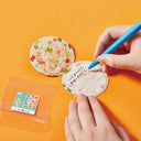 A close-up of hands holding a blue marker and writes on a round paper card printed with shrimp and vegetable patterns.