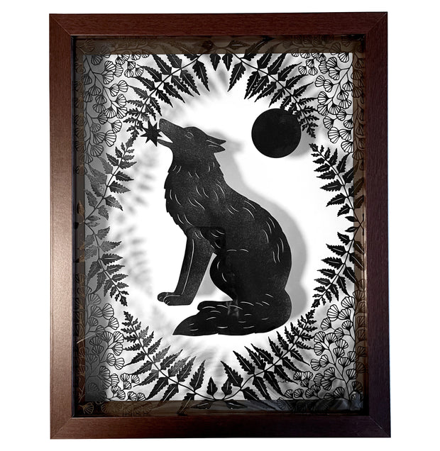 Black paper cutting in a shadowbox style frame with a glass back. A wolf sits and holds a star in its mouth, it is framed by delicately cut fern leaves and flowers and a full moon.