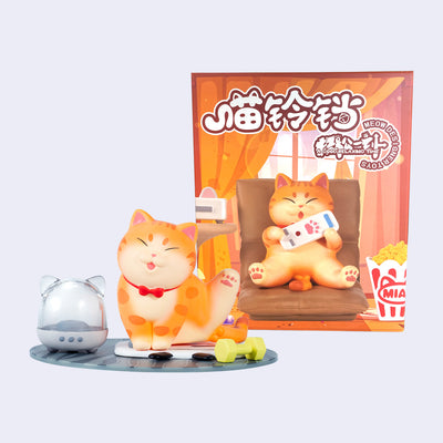 Plastic figure of a orange tabby cat, sitting on a mat and raising its leg as if doing yoga. Next to it is a small dumbbell and a humidifier like object, shaped as a cat head. It stands in front of its product packaging.