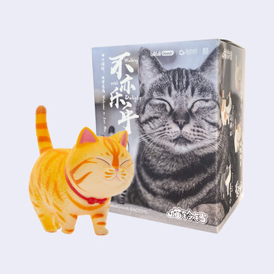 Orange tabby cat figurine, cartoon styled with a red bowtie around its neck and a happy closed eye expression. It walks on all 4s with with tail sticking straight up.