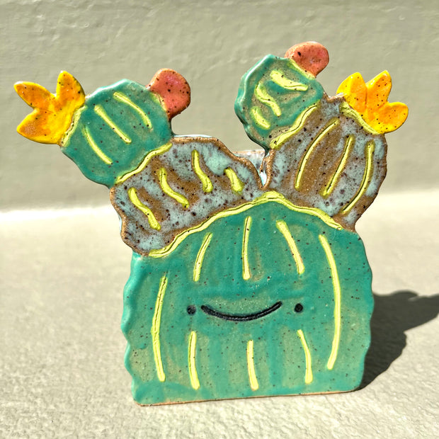 Flat ceramic sculpture of a cactus with barrel like appendages atop its head. It is simplistic in form and has a smiling cartoon face. It has several flowers blooming atop of it.