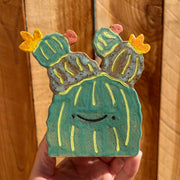 Flat ceramic sculpture of a cactus with barrel like appendages atop its head. It is simplistic in form and has a smiling cartoon face. It has several flowers blooming atop of it.