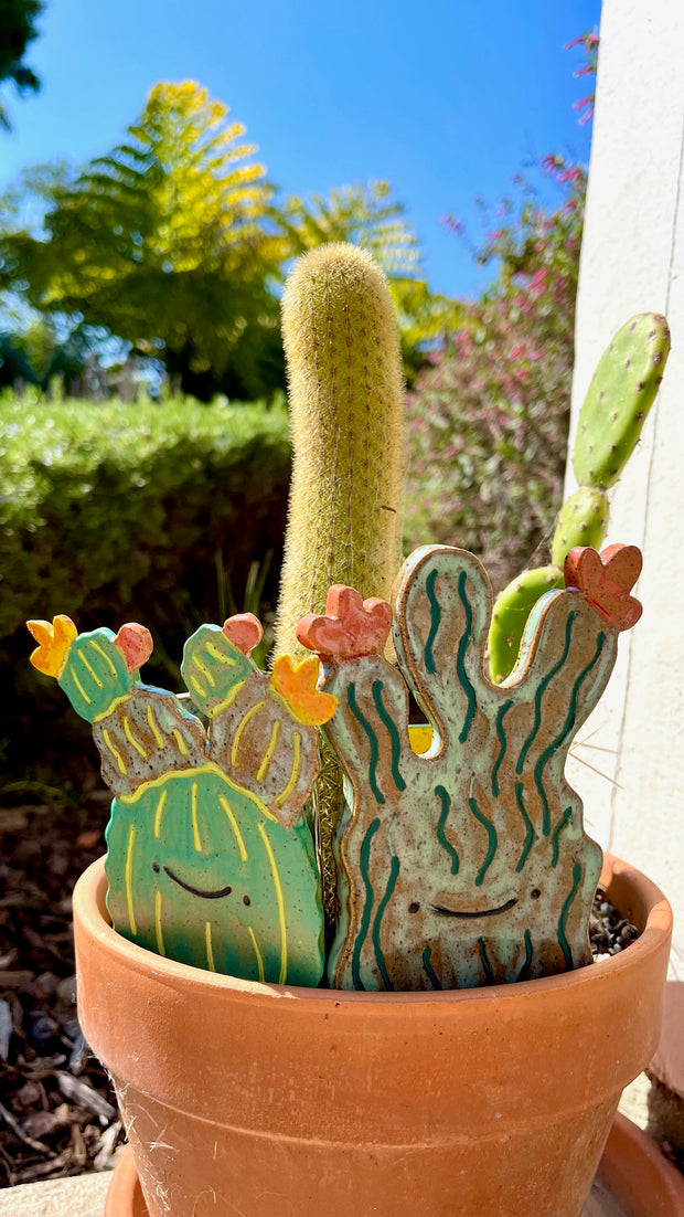 Ceramic sculpture of 2 flat cartoon style cacti, both with smiling faces and simplistic features. Each have flowers blooming from them.