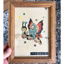 Framed illustration in a traditional tattoo style of a fairy butterfly riding atop of a caterpillar with reins. 