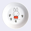 White shallow bowl with a graphic of Miffy, wearing an orange dress and standing with her hands behind her back. Small white flowers with yellow centers are around her feet.