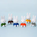Six small rabbit figurines sitting in a row, each with white ears and body and wearing different colored outfits: green, brown, blue, orange, yellow, and gray, on a light blue background.