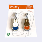 2 die cut metal charms, one of Miffy in a blue dress and one of Miffy's friend Melanie, in her orange dress. Each are attached to their own clasp on a white backing card. Shown at an angle to display sheen.