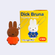 A brown bunny version of Miffy, in a bright reddish orange dress shirt, standing with a keychain attached. It stands in front of a yellow blind box packaging.