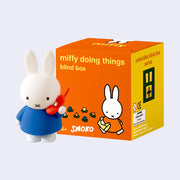 Figurine of Miffy standing in a blue dress and talking on an orange phone. She stands in front of her orange product packaging box.